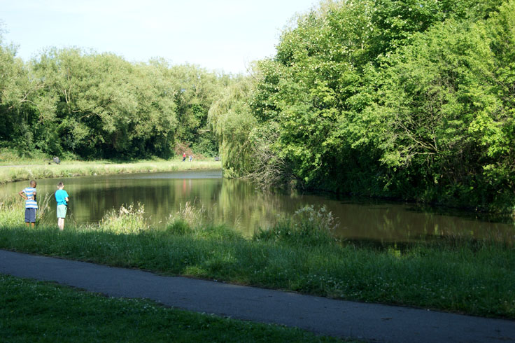 Two boys standing by a lake, with some people walking along the other side.